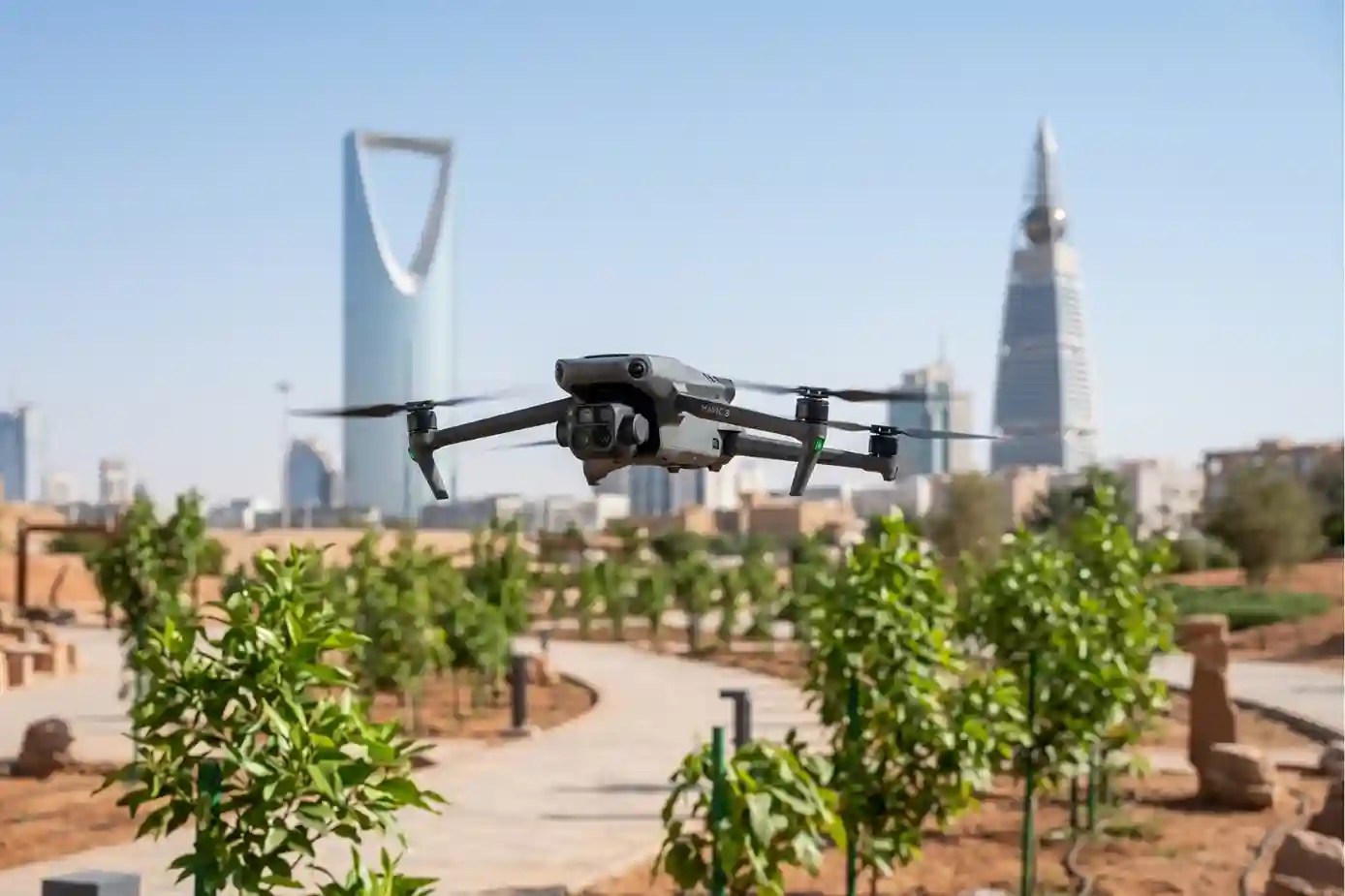 A compact DJI Mavic 3 Multispectral drone hovering over a newly planted green park path in Riyadh during a bright summer day.