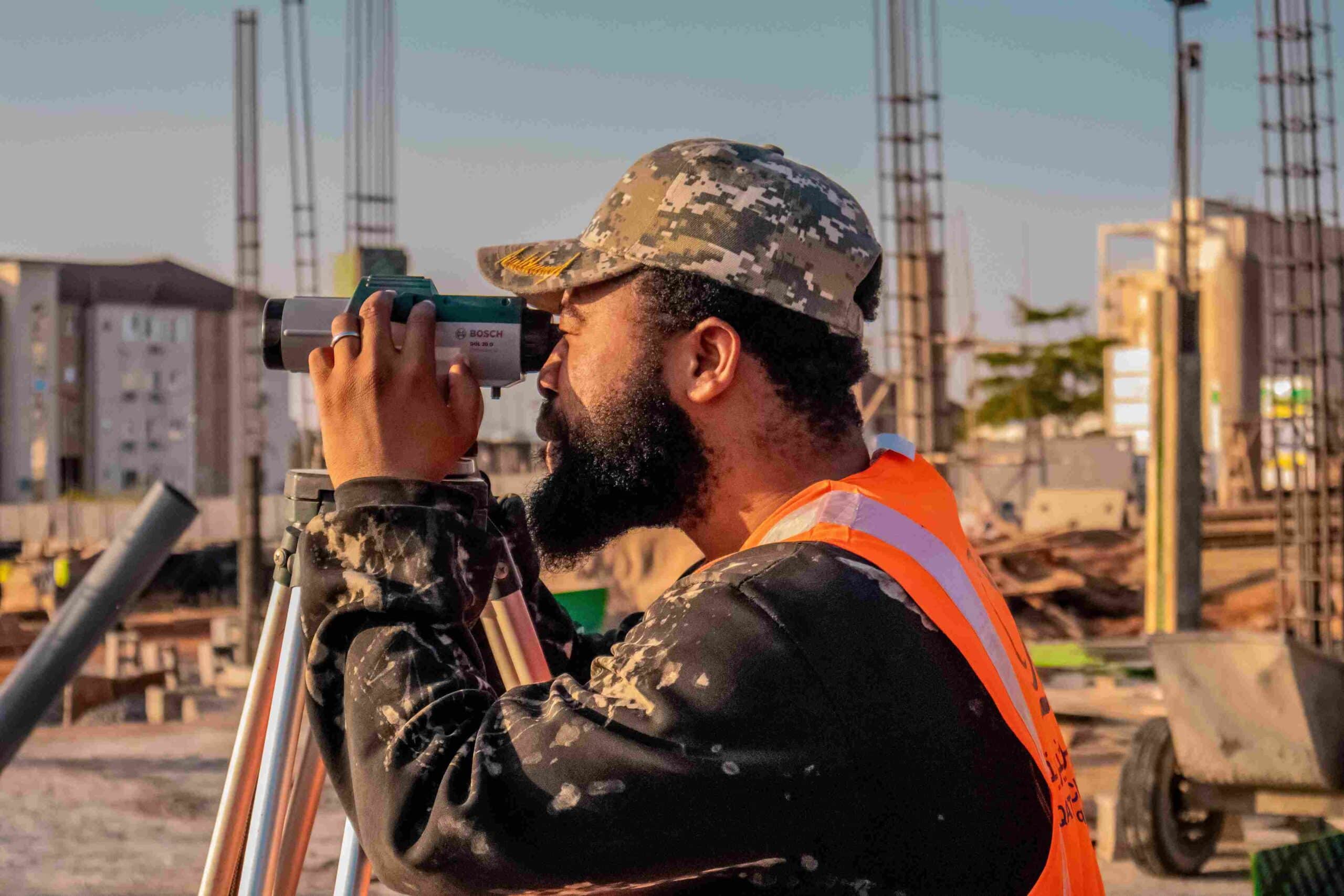 A professional surveyor with a beard, wearing a camouflage cap and high-visibility orange safety vest, looking through a tripod-mounted Bosch optical level to map ground elevations at an active infrastructure construction site.