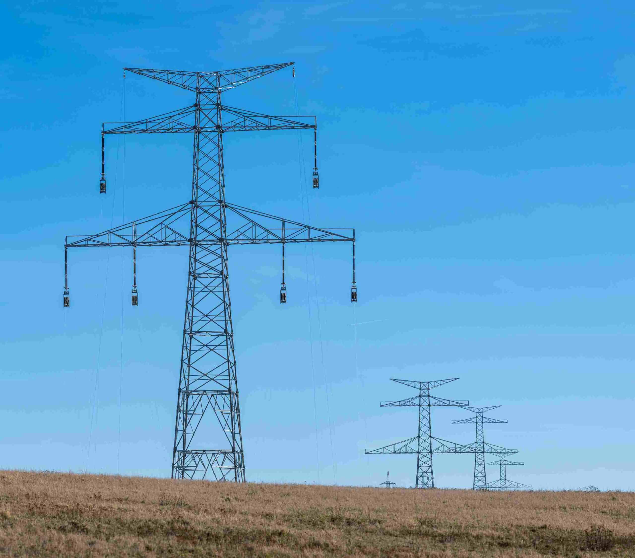 A large steel lattice high-voltage transmission tower stands on a dry, rolling hill, with a row of successive power towers extending into the distance along a utility corridor under a clear blue sky as shown in Transmission towers.jpg.