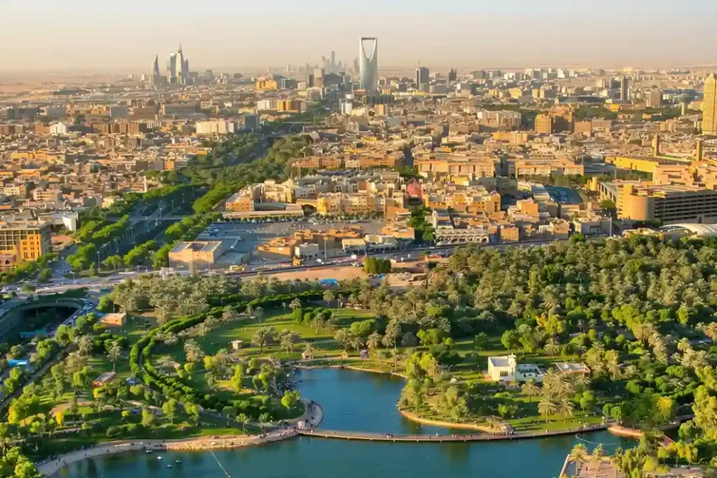 A wide aerial panoramic shot showing a massive green municipal park development project seamlessly integrated with surrounding modern urban neighborhoods in Riyadh under a bright clear sky.