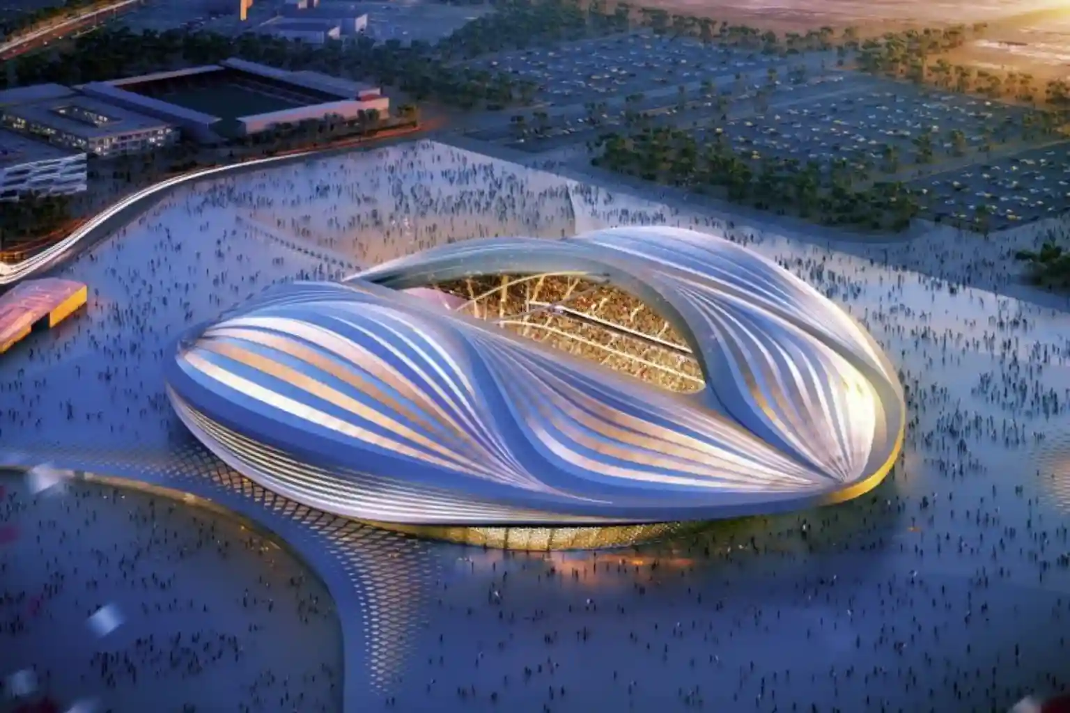 High-angle wide aerial photograph of a massive sports stadium undergoing heavy reconstruction, featuring exposed stadium seating tiers and large geometric roof canopy frames under a clear blue sky.