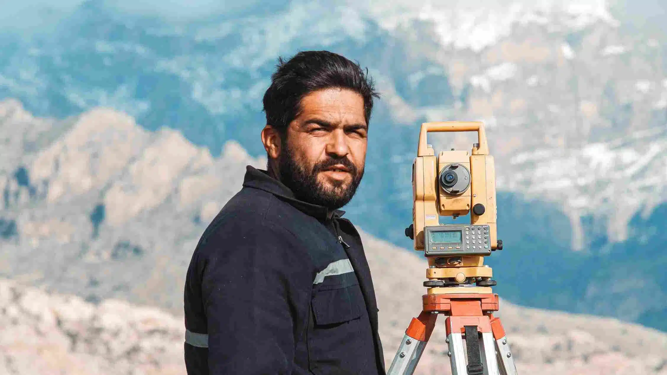 A close-up photograph of a bearded male surveyor standing next to a yellow optical total station mounted on a tripod, with rugged, hazy mountains in the background.