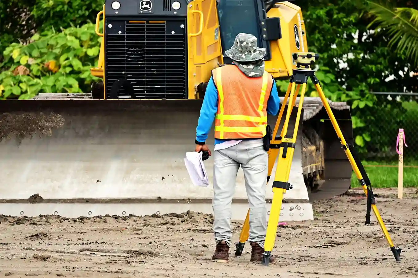 A land surveyor in an orange safety vest and sun hat holding survey documents while standing beside a yellow tripod and surveying instrument with heavy construction machinery in the background.