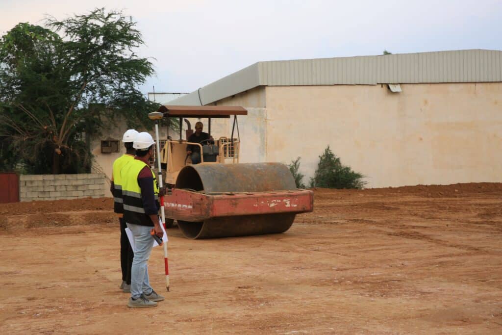Two land surveyors using a GNSS DGPS survey pole and field controller to take elevation measurements at an active construction site.
