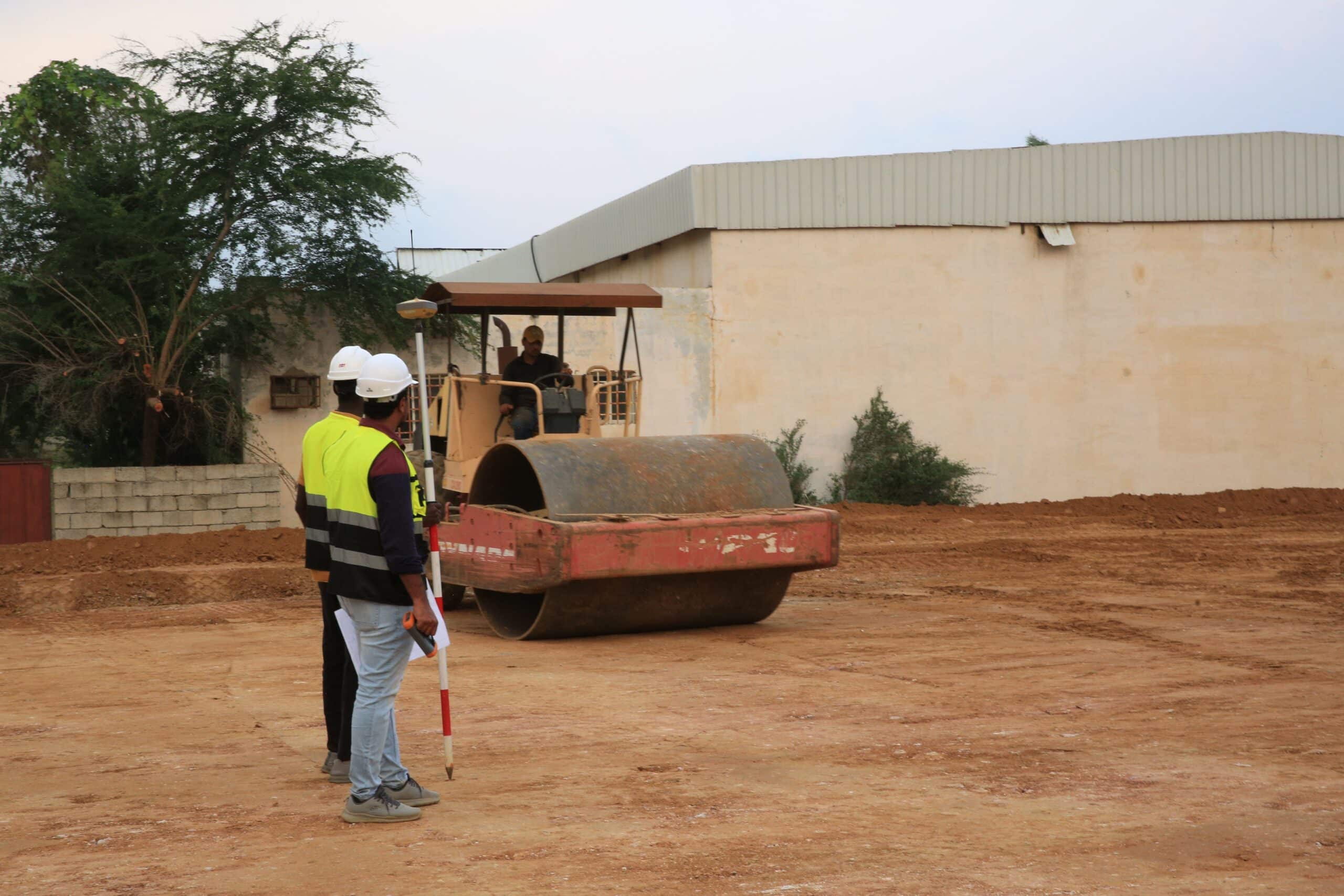Two land surveyors using a GNSS DGPS survey pole and field controller to take elevation measurements at an active construction site.