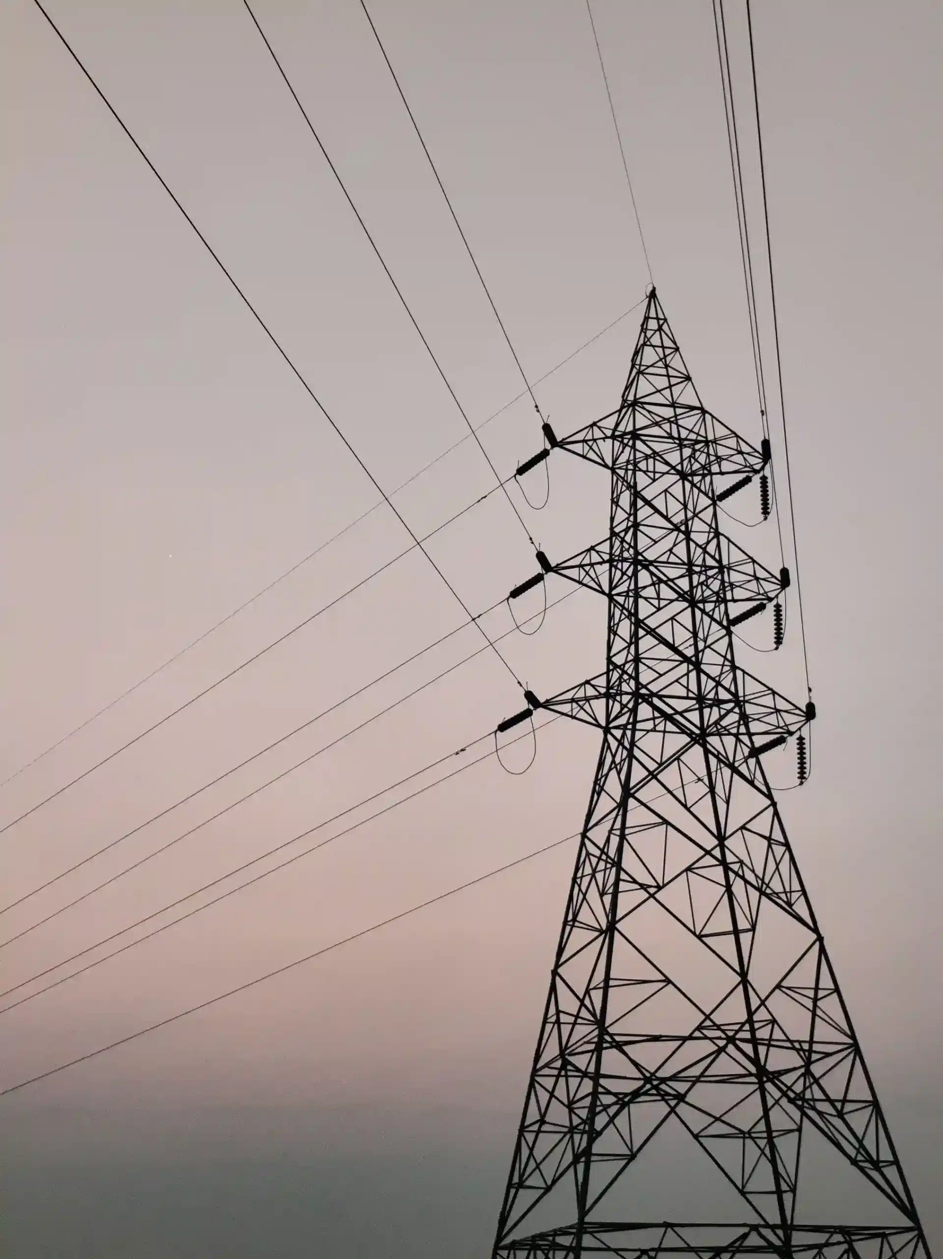 High-voltage steel lattice transmission tower supporting overhead power lines along a utility transmission corridor against a twilight sky.