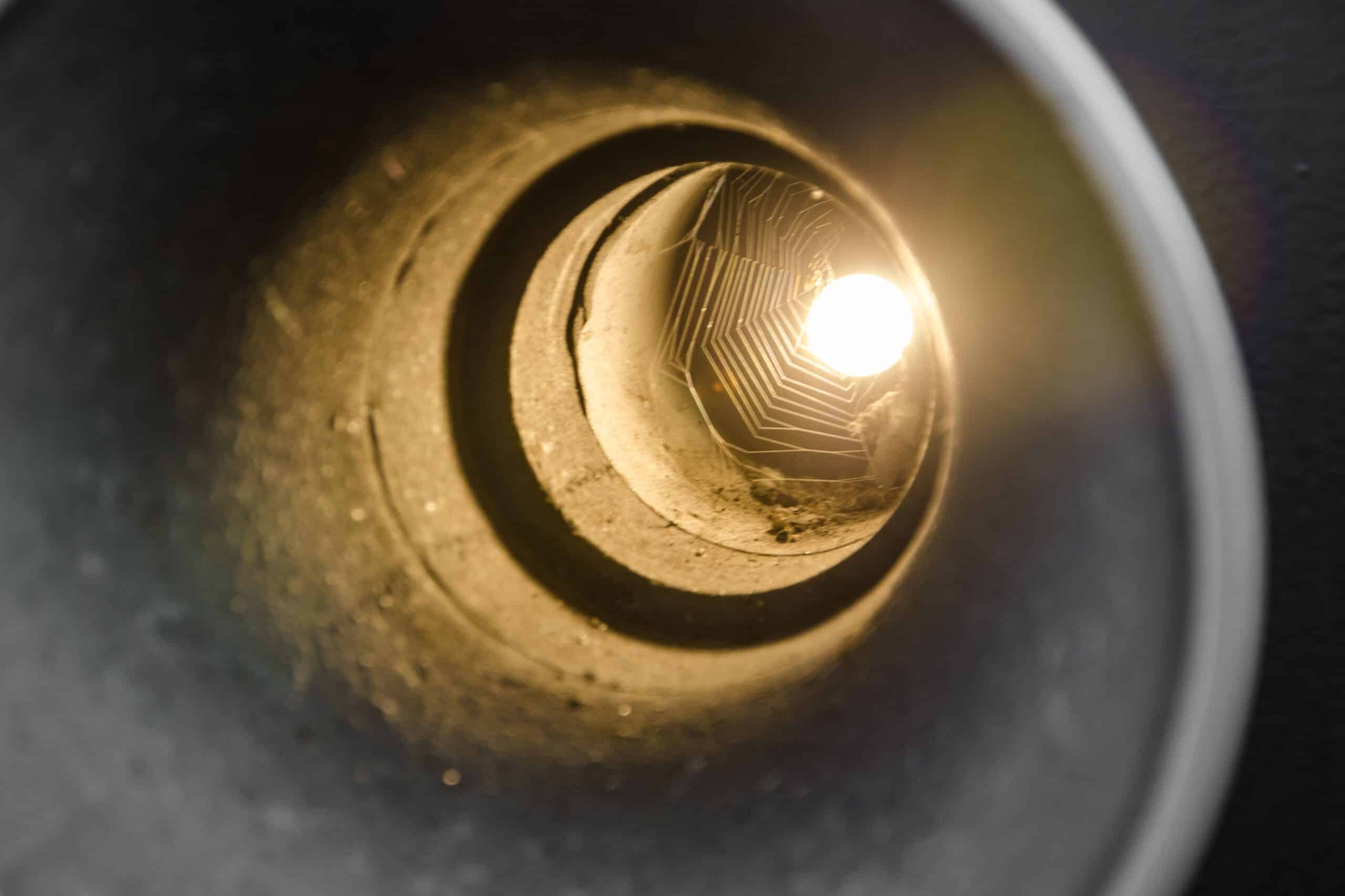 Interior view looking down a narrow circular sewer pipe illuminated by inspection lighting showing pipe wall conditions and internal obstructions.